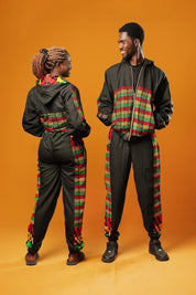 Woman and man standing against an orange background wearing black tracksuit trousers with colourful Kente African print panels down the sides. The trousers have a relaxed fit with elastic cuffs at the ankles and a drawstring waist. They both pair them with matching black jackets featuring Kente trim.