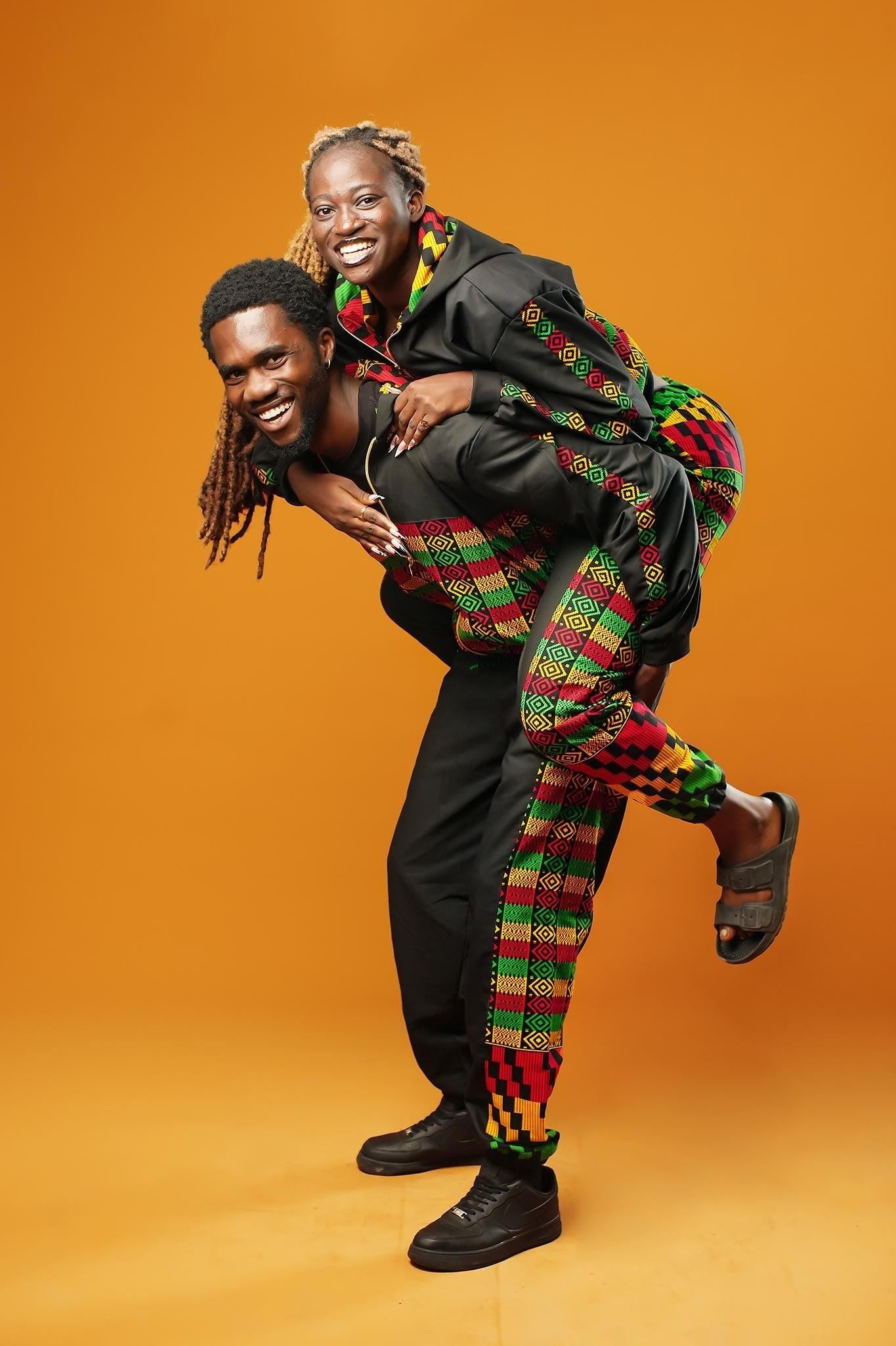 Woman and man standing against an orange background wearing black tracksuit trousers with colourful Kente African print panels down the sides. The trousers have a relaxed fit with elastic cuffs at the ankles and a drawstring waist. They both pair them with matching black jackets featuring Kente trim.