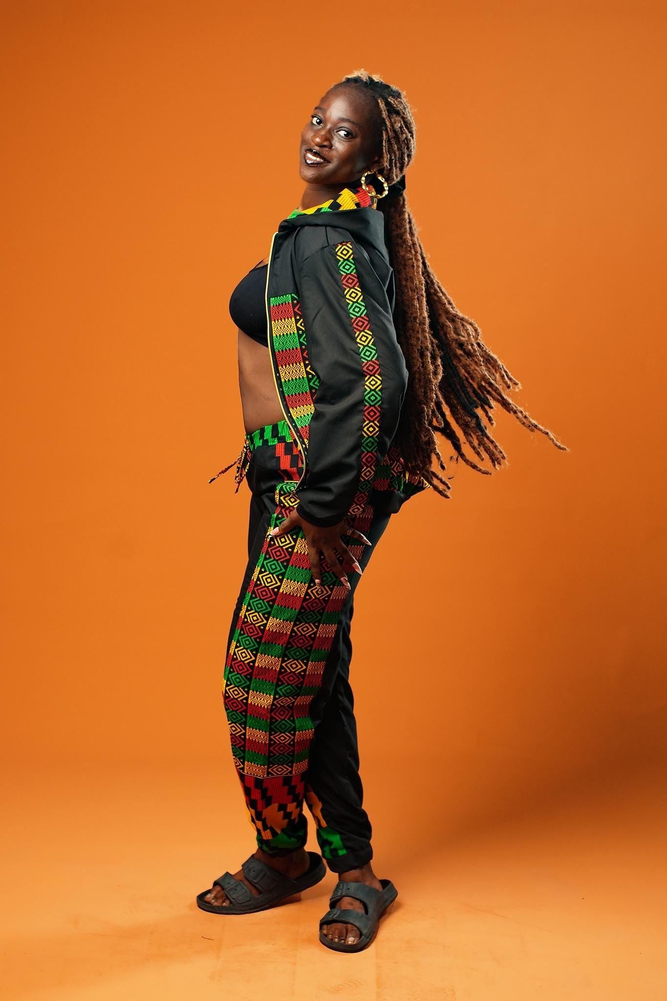 Woman standing against an orange background wearing black tracksuit trousers with colourful Kente African print panels down the sides. The trousers have a relaxed fit with elastic cuffs at the ankles and a drawstring waist. She pairs them with a matching black jacket featuring Kente trim and a black crop top.