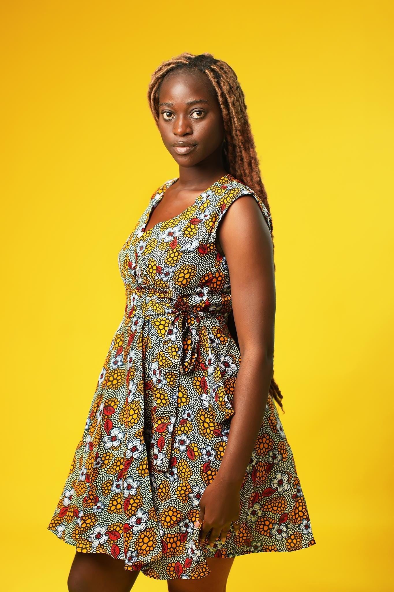 Close up of a woman wearing a sleeveless African print wrap dress made from colourful ankara fabric with yellow, red, and white floral patterns. The dress features a V-neckline and tie waist, and falls above the knees.