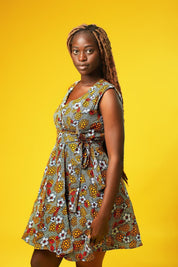 Close up of a woman wearing a sleeveless African print wrap dress made from colourful ankara fabric with yellow, red, and white floral patterns. The dress features a V-neckline and tie waist, and falls above the knees.