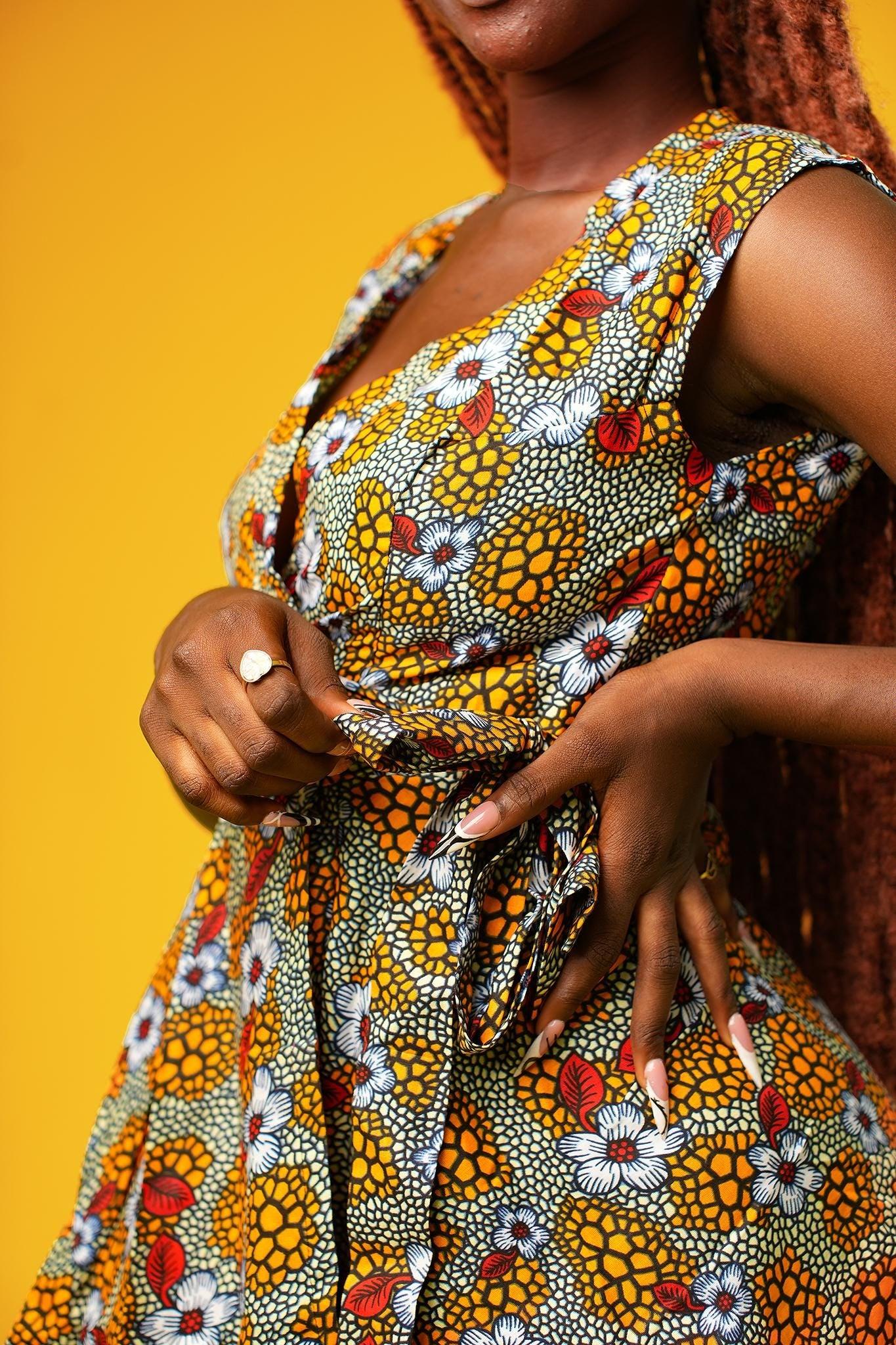 Woman wearing a sleeveless African print wrap dress made from colourful ankara fabric with yellow, red, and white floral patterns. The dress features a V-neckline and tie waist, and falls above the knees.