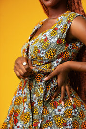 Woman wearing a sleeveless African print wrap dress made from colourful ankara fabric with yellow, red, and white floral patterns. The dress features a V-neckline and tie waist, and falls above the knees.
