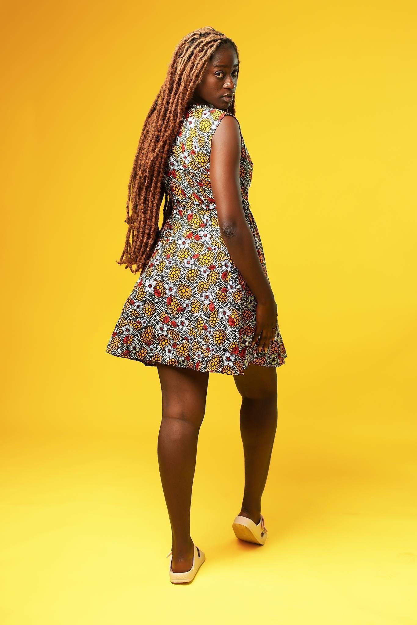 Woman wearing a sleeveless African print wrap dress made from colourful ankara fabric with yellow, red, and white floral patterns. The dress features a V-neckline and tie waist, and falls above the knees.